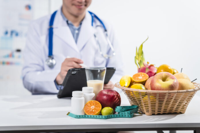 A male nutritionist working at a hospital desk, holding an apple and mixed fruit, offering care, health advice, weight loss guidance, and eating tips online for patients seeking nutrition support