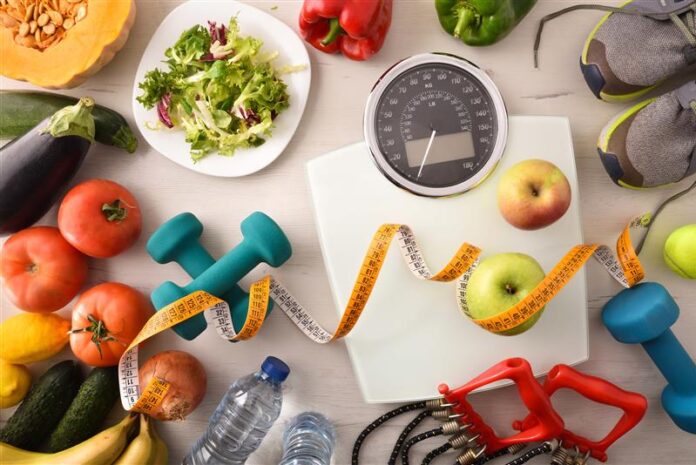 Una bilancia, degli strumenti per far esercizio come pesi ed elastici e cibo tipico della dieta mediterranea
