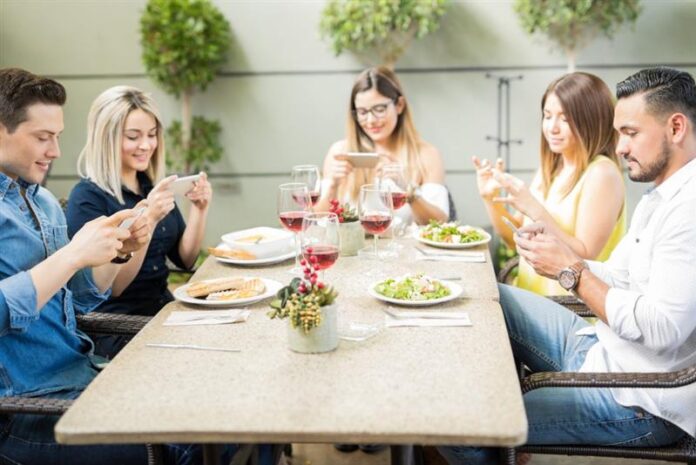 AdobeStock_131453285 Persone che mangiano a un tavolo con il telefono in mano